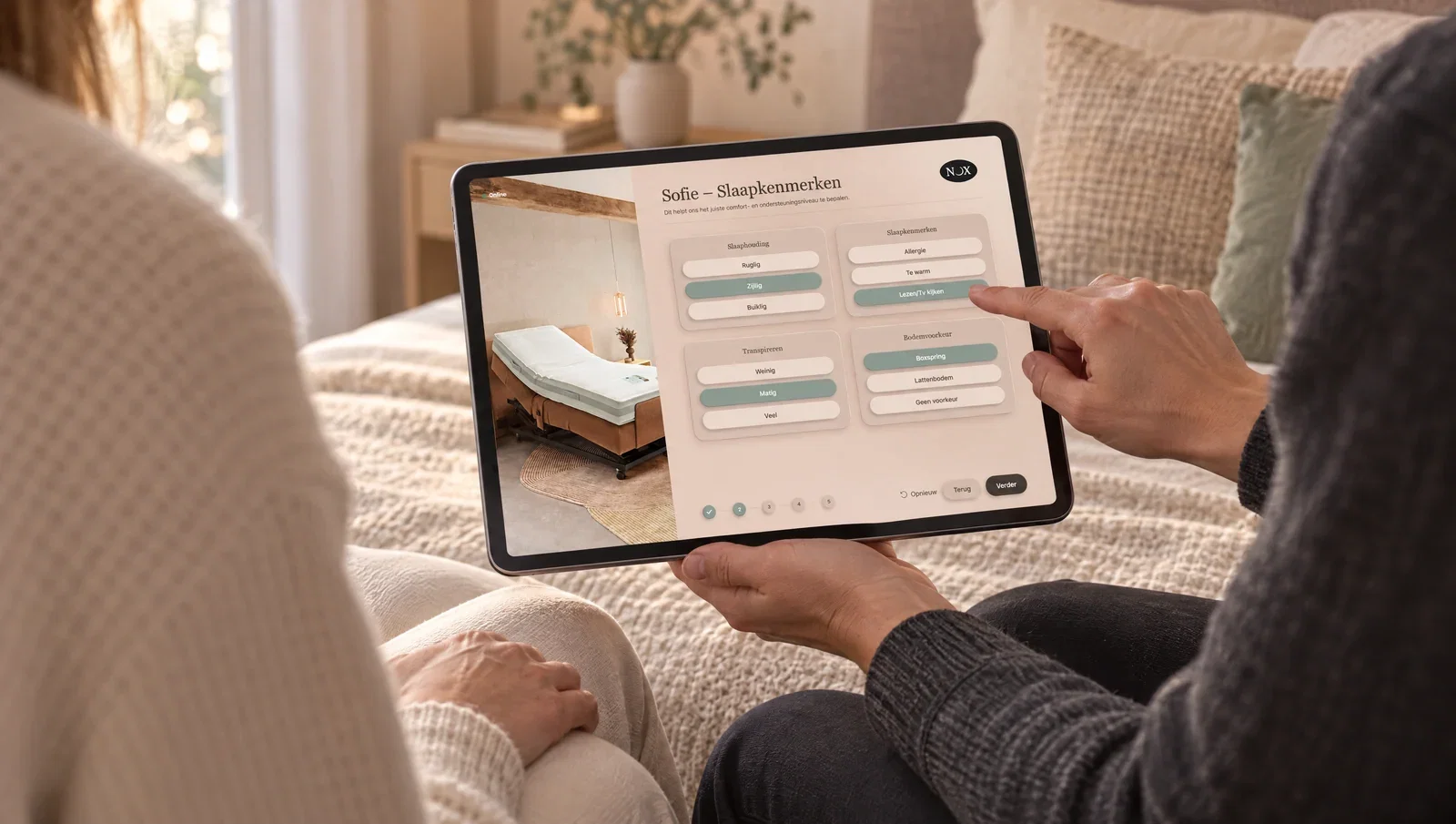A salesperson and a customer on the edge of a bed, looking at the NOX configurator on a tablet together