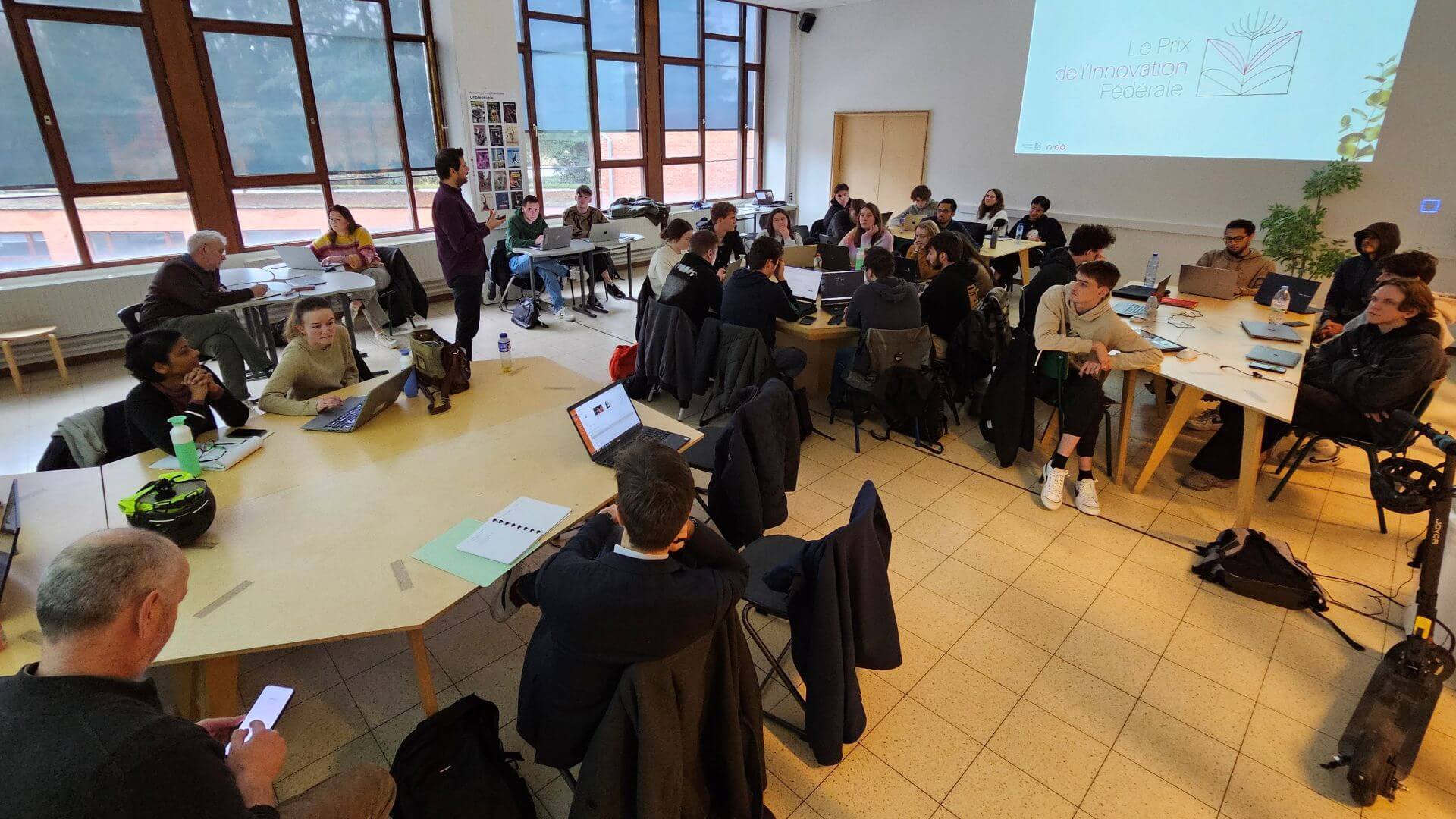 Groep studenten zit rondom tafels in een klaslokaal met laptops en notities, terwijl iemand vooraan staat en spreekt.