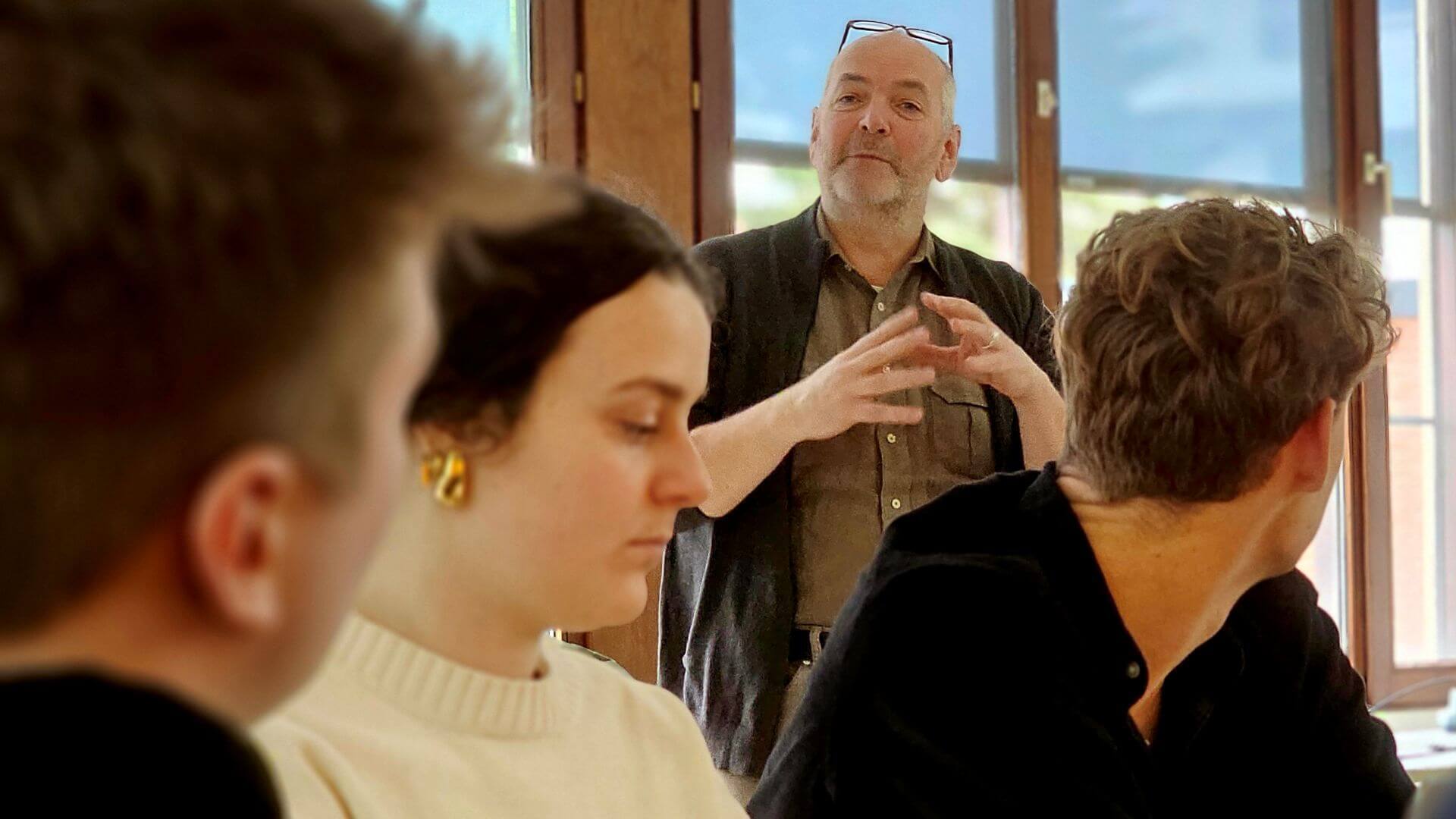 Docent staat vooraan in een leslokaal en legt iets uit aan een kleine groep studentendie aan een tafel zitten en aandachtig luisteren.